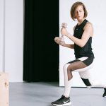A young woman performing lunges with dumbbells in a modern indoor gym setting.
