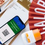 Flat lay of travel essentials featuring a digital green pass, passport, and other travel items on a table.